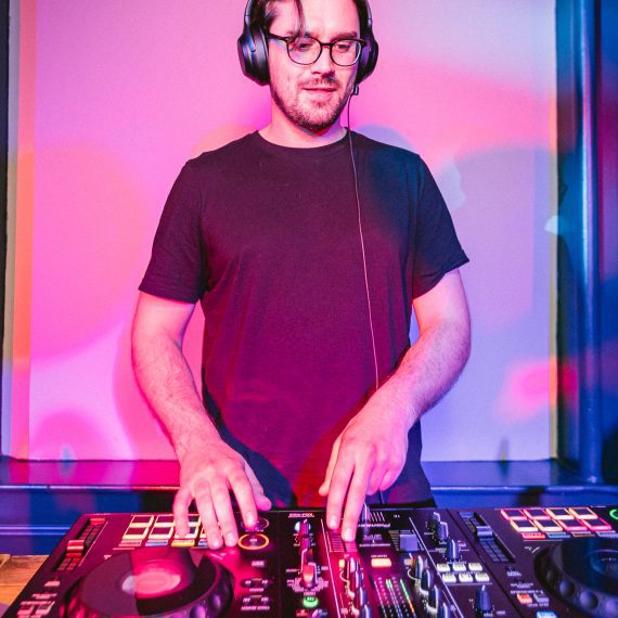 Concentrated wedding DJ for hire against a backdrop of vibrant pink and blue lights at a lively wedding reception.